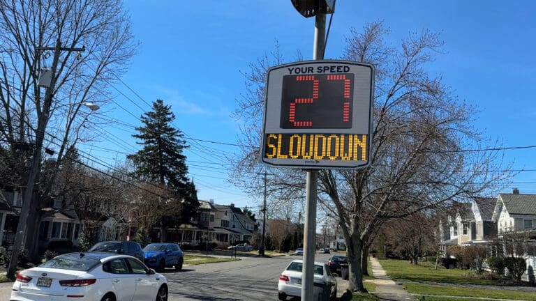 The image shows a speed monitoring sign on a residential street. The sign displays "Your Speed" in small letters at the top, with "27" in large red numbers, and "SLOWDOWN" in yellow letters below the speed