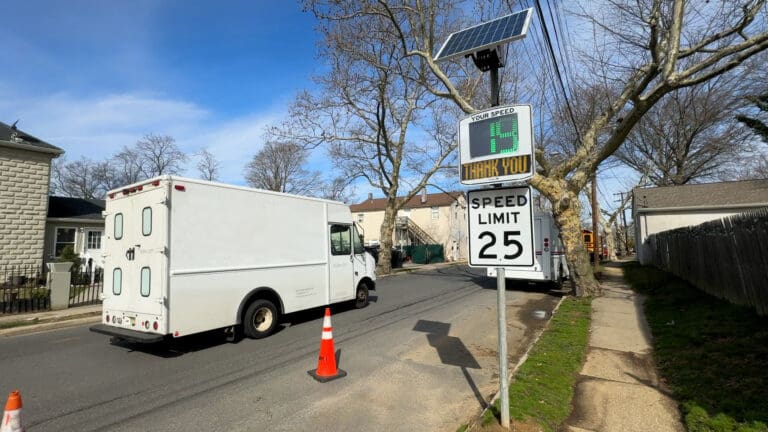 White truck passing by a solar-powered speed feedback sign displaying "Thank you" and a speed of 14 mph, with a sign indicating a speed limit of 25 mph on a residential street.